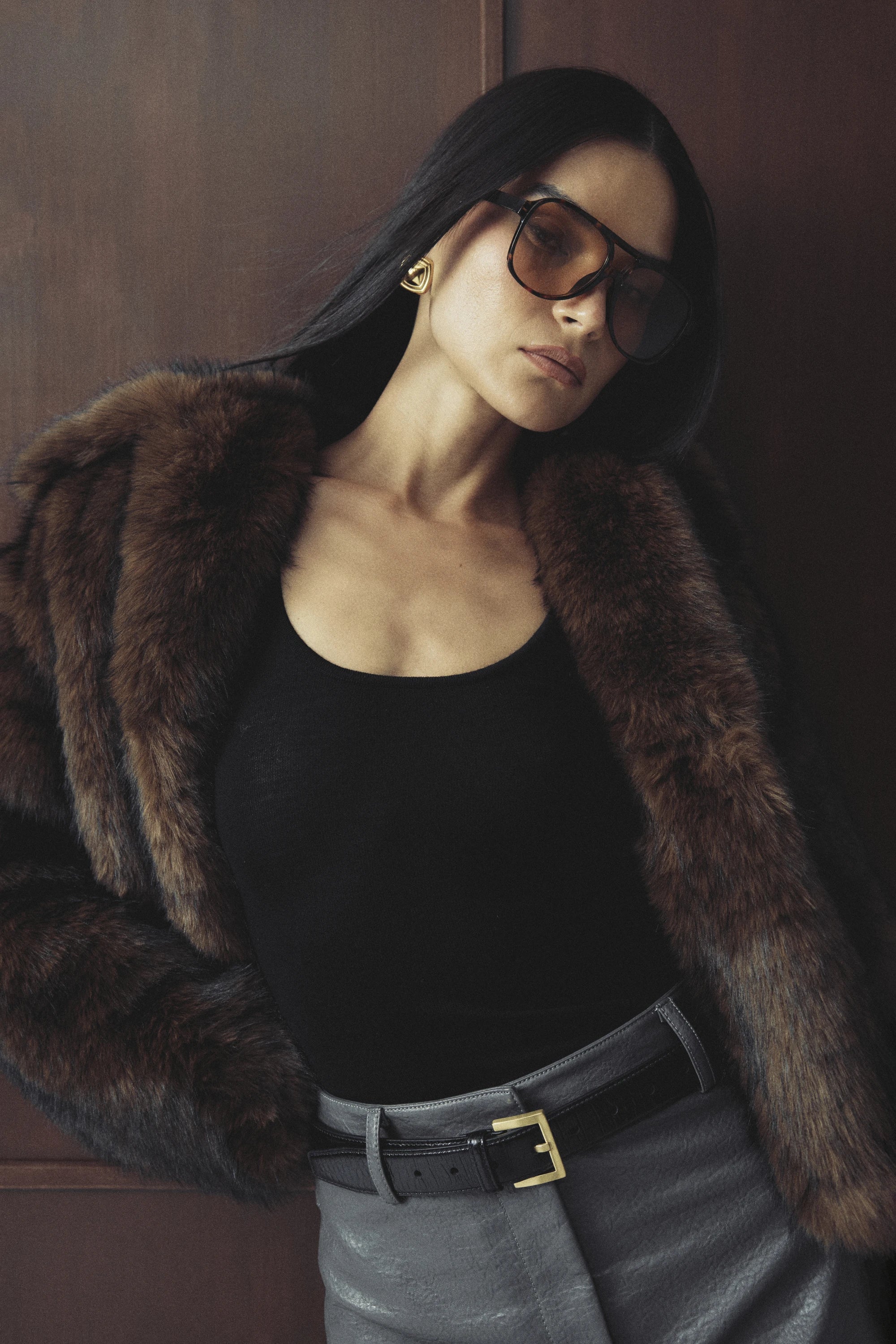 Woman wearing a brown fur coat, black top, and gray pants against a wooden wall.